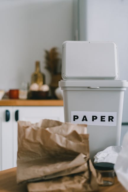 A person is shown placing a crumpled brown paper bag into a white waste bin with a removable lid, positioned indoors on a wooden surface. The individual's arm and hand are visible, with the hand gripp